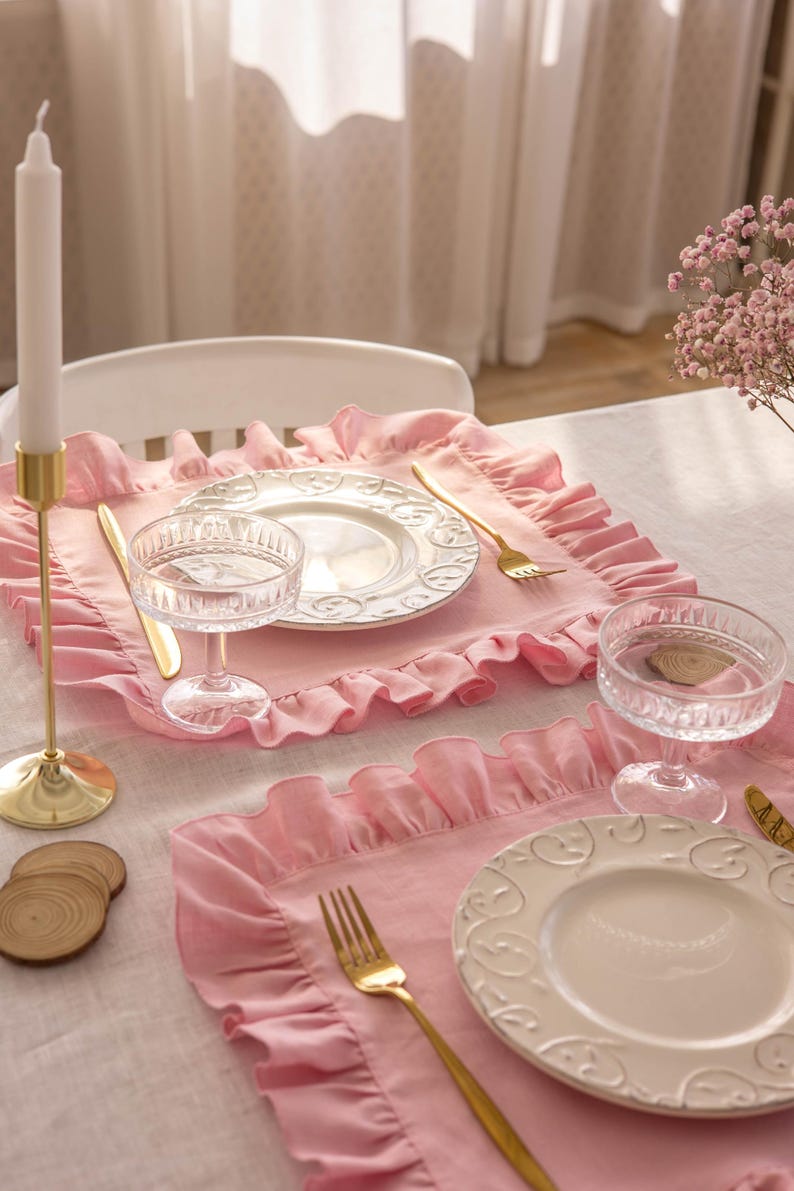 Puede incluir: Dos manteles individuales de color rosa con volantes, con platos blancos y cubiertos dorados. Los manteles individuales est&aacute;n sobre un mantel de lino blanco.