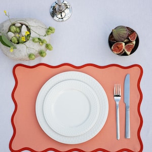 May include: A table setting featuring a coral placemat with a red scalloped border. Two white plates, a fork, and a knife are arranged on the mat. A small bowl of figs and a decorative stone bowl with greenery are also visible.