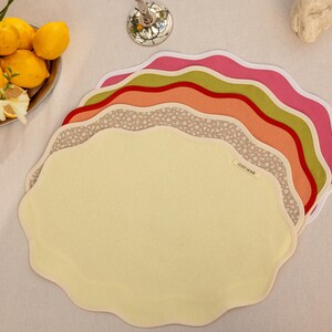 Op de afbeelding: Een stapel placemats met geschulpte randen in verschillende kleuren, waaronder geel, roze, oranje, groen en een bloemenprint. De placemats zijn gerangschikt op een wit oppervlak, met een kom met citroenen op de achtergrond. De tekst "COZY HOME" is zichtbaar op een van de placemats.