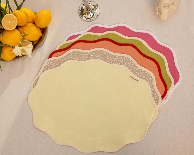 Op de afbeelding: Een stapel placemats met geschulpte randen in verschillende kleuren, waaronder geel, roze, oranje, groen en een bloemenprint. De placemats zijn gerangschikt op een wit oppervlak, met een kom met citroenen op de achtergrond. De tekst "COZY HOME" is zichtbaar op een van de placemats.