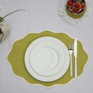 Op de afbeelding: Een tafelsetting met een groene placemat met een geschulpte rand, twee witte borden, een vork en een mes. Een kleine kom met vijgen staat op een kanten kleedje. De tafel is bedekt met een wit tafelkleed.