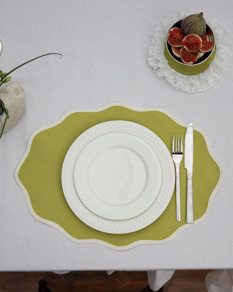 Op de afbeelding: Een tafelsetting met een groene placemat met een geschulpte rand, twee witte borden, een vork en een mes. Een kleine kom met vijgen staat op een kanten kleedje. De tafel is bedekt met een wit tafelkleed.