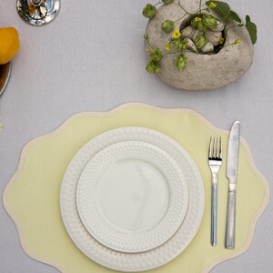 Op de afbeelding: Een tafelsetting met een lichtgele placemat met een geschulpte rand, twee witte borden met textuur, een vork en een mes. Een decoratieve stenen kom met groen en kleine gele bloemen is aanwezig. Een zilveren kandelaar en een citroen zijn ook zichtbaar.