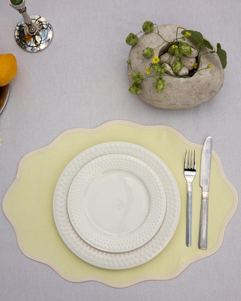 Op de afbeelding: Een tafelsetting met een lichtgele placemat met een geschulpte rand, twee witte borden met textuur, een vork en een mes. Een decoratieve stenen kom met groen en kleine gele bloemen is aanwezig. Een zilveren kandelaar en een citroen zijn ook zichtbaar.