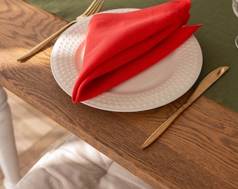 Puede incluir: Una servilleta de lino roja doblada en forma de triángulo se coloca sobre un plato blanco con un patrón de puntos. El plato está sobre una mesa de madera con un mantel individual de lino verde. Un tenedor y un cuchillo dorados están sobre la mesa.