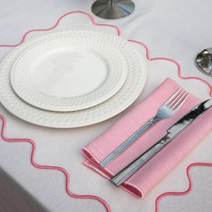 Peut inclure: Une table dressée avec des assiettes blanches, un set de table rose avec une fourchette et un couteau, et une bordure rose décorative. Les assiettes ont un bord texturé. L'ensemble est sur une nappe blanche.