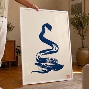 Puede incluir: Una impresión de arte mural vertical enmarcada que presenta una ilustración de una serpiente en azul oscuro, estilo pincelada, sobre un fondo crema claro. La obra incluye un pequeño sello cuadrado rojo en la esquina inferior derecha, todo en un marco blanco fino.
