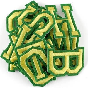 May include: A collection of green and gold embroidered letter patches. The letters, in a classic collegiate style, have a gold centre and a green border. These patches are likely for personalising clothing or accessories.