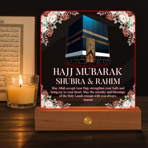May include: An illuminated sign with the text "HAJJ MUBARAK SHUBRA & RAHIM" and an image of the Kaaba. The sign is set on a wooden base and has floral decorations. A lit candle and a book are also visible.