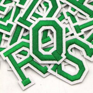 May include: Close-up of green embroidered letters with white borders. The letters are in a sans-serif font and are scattered across the image. The letters are made of a soft, textured material.