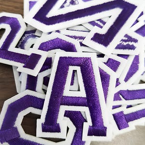 May include: A collection of purple embroidered varsity letters with white edging. The letters, in a bold, block design, have a textured appearance. They are scattered on a wooden surface.
