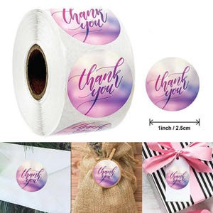 May include: A roll of round stickers with a watercolour background in shades of purple and pink. Each sticker has the words "thank you" in elegant cursive script. One sticker is 2.5cm in diameter.