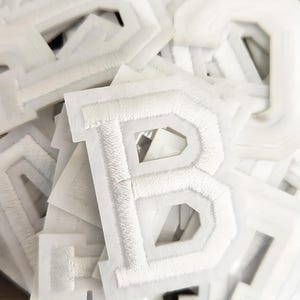 May include: A collection of white, embroidered letter patches. The letters, in a serif font, have a fluffy, textured look. The patches are on a white backing, creating a layered effect. Ideal for customising clothing.