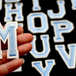 May include: A close-up of a light blue embroidered letter "M" patch with a white border, held in a hand. Other light blue embroidered letters are in the background against a black surface. The patches are likely for clothing or crafts.