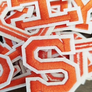 May include: A close-up shot of a collection of orange embroidered varsity letter patches with white borders. The letters are in a bold, block style, with a textured, stitched appearance. These patches are likely for customising clothing.