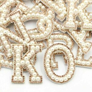 May include: Close-up of various white, pearl-studded letters with a gold-coloured rhinestone border. The letters are scattered, showcasing the intricate details of the embellishments. The letters are likely for crafting or decorative purposes.