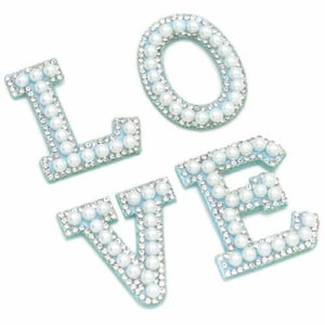 May include: Four individual letters spelling "LOVE". Each letter is light blue and outlined with small clear rhinestones. The letters are further embellished with white faux pearls, creating a decorative and elegant design.