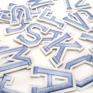 May include: A selection of embroidered fabric letters in various fonts. The letters are light blue with a white border, scattered on a white surface. These are likely patches for clothing or craft projects.