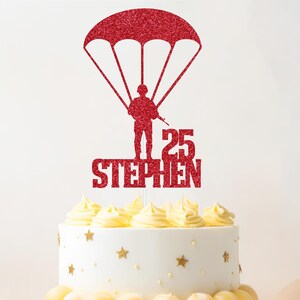 May include: A red glitter cake topper depicting a soldier under a parachute, with the name "STEPHEN" and the number "25". The topper sits atop a white cake decorated with yellow icing and gold star decorations.