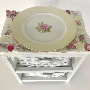 May include: A decorative white table with two wicker drawers, topped with a mosaic of floral tiles and a pale yellow plate. The plate features a pink rose design and gold trim. Embellished with pink brooches.