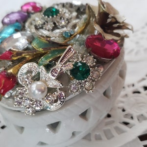 May include: A white ceramic trinket box decorated with colorful rhinestones, faux pearls, and metallic accents. The box is shaped like a heart and has a delicate, intricate design.