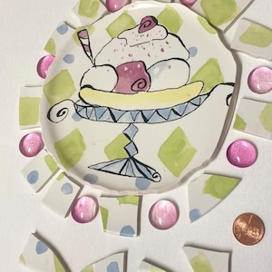 May include: A whimsical, hand-painted ceramic banana split dish with a scoop of ice cream, cherries, and a spoon. The dish is surrounded by broken ceramic pieces and pink glass gems. A penny is included for scale.