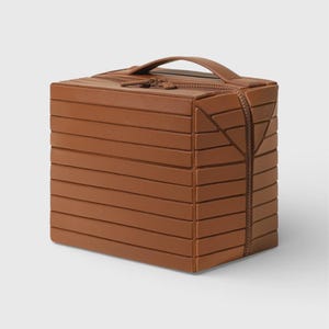 May include: A brown, box-shaped bag with a handle and zip closure. The bag is made from horizontal, layered panels, with a zip running along the top and down one side. The design features geometric accents and a sturdy handle.