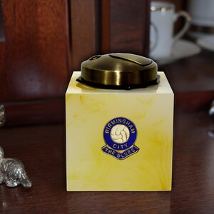 May include: A yellow and white marble cigarette lighter with a brass top. The lighter features a blue and gold Birmingham City Football Club logo with the text "Birmingham City The Blues".