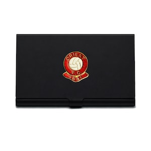 May include: A black business card holder with a red, white, and gold emblem. The emblem displays a football and the text "ORIENT F.C. 1881". The card holder is rectangular with a small recess on the bottom edge.
