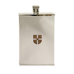 May include: A rectangular silver-toned metal flask with a polished finish and a screw-top cap. The centre of the flask features a small, colourful heraldic shield emblem displaying four lions and a central crown on a white and red background.