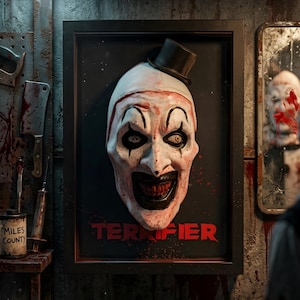 Puede incluir: Un cuadro decorativo con una máscara de payaso blanca en relieve, un sombrero de copa negro, maquillaje oscuro en los ojos y una sonrisa amplia. El texto TERRIFIER aparece en rojo en la parte inferior. El marco negro está sobre una pared oscura.