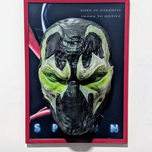 Könnte beinhalten: Eine gerahmte Spawn-Maske mit rotem Rand. Die Maske ist schwarz, grün und grau, mit leuchtend grünen Augen. Der Hintergrund zeigt das Spawn-Logo und den Text "BORN IN DARKNESS, SWORN TO JUSTICE."