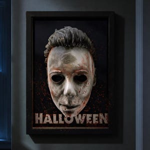Könnte beinhalten: Ein gerahmtes dreidimensionales Wandkunstwerk mit einer blassen, verwitterten Maske mit dunklen Augenhöhlen und roten Spritzern. Das Wort HALLOWEEN ist in fetten, abgenutzten Großbuchstaben am unteren Rand des schwarzen rechteckigen Rahmens gedruckt.