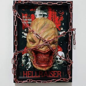 May include: A framed Hellraiser artwork featuring a grotesque, textured face with an open mouth and visible teeth, wrapped in red-painted chains. The background is a movie poster with the title "Hellraiser" and other characters.
