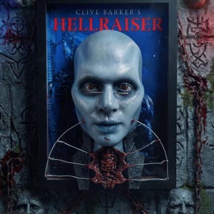 May include: A framed 3D wall art piece featuring a pale blue humanoid head with metal pins in the cheeks and eyes, set against a dark blue background. The text CLIVE BARKER'S HELLRAISER is printed in red above the head, with a decorative wire structure.