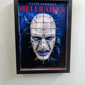 Peut inclure: Œuvre encadrée représentant le personnage de Pinhead de Hellraiser de Clive Barker. L'image montre le visage de Pinhead, avec des clous et des lignes rouges sur sa tête. Le fond est bleu foncé et le cadre est noir.