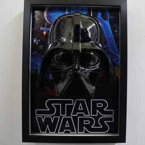 Op de afbeelding: Ingelijste Star Wars-kunst met een glanzend zwart Darth Vader-masker tegen een ruimteachtergrond. Het Star Wars-logo staat in witte blokletters onderaan het frame. Het frame is zwart.