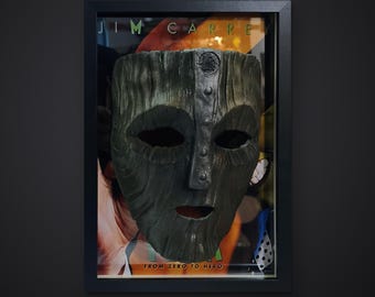 Può includere: Una maschera testurizzata verde scuro con una cresta centrale verticale e borchie circolari, esposta in una cornice nera sopra un poster con il testo JIM CARREY e FROM ZERO TO HERO. La maschera presenta orbite profonde e una piccola bocca.