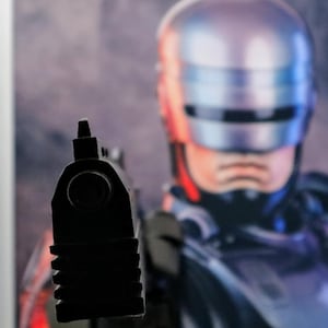 Pu&ograve; includere: Immagine ravvicinata di una pistola nera puntata direttamente verso l'osservatore. Sullo sfondo, un'immagine sfocata di un elmo metallico e della parte superiore del corpo di una figura, probabilmente di fantascienza, &egrave; visibile.