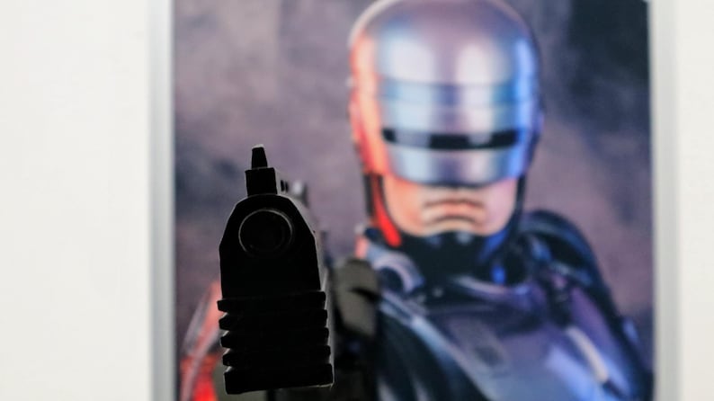 Pu&ograve; includere: Immagine ravvicinata di una pistola nera puntata direttamente verso l'osservatore. Sullo sfondo, un'immagine sfocata di un elmo metallico e della parte superiore del corpo di una figura, probabilmente di fantascienza, &egrave; visibile.