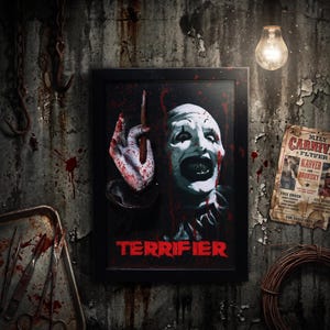 Pode incluir: Uma peça de arte de parede 3D emoldurada apresentando Art, o palhaço de Terrifier, com rosto branco, olhos pretos e dentes afiados. O palhaço segura uma ferramenta manchada de sangue, com salpicos vermelhos e a palavra Terrifier em vermelho.