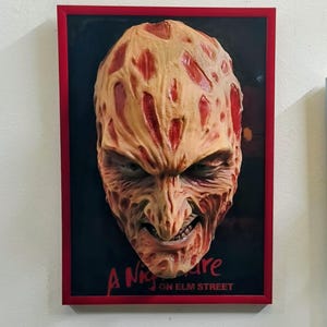 May include: A framed artwork featuring a detailed, textured mask of a character with a scarred face, set against a black background. The frame is red, and the text "A Nightmare on Elm Street" is visible at the bottom.