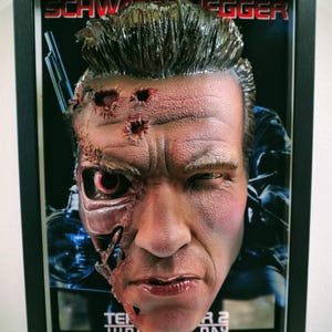 Peut inclure: Œuvre d'art 3D encadrée représentant un buste de cyborg détaillé avec des composants mécaniques exposés et des dommages de combat. L'œuvre est placée sur un fond d'affiche de film avec le nom "SCHWARZENEGGER" visible. Le cadre est noir.