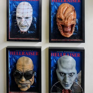 May include: Four framed wall displays featuring 3D character masks from Clive Barker's Hellraiser. Each black frame contains a blue background with the text CLIVE BARKER'S HELLRAISER and a unique sculpted mask including Pinhead and other creatures.