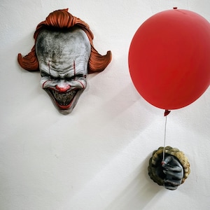 Può includere: Una scultura da parete raffigurante un volto di clown pallido e sorridente con denti affilati e capelli arancioni, abbinata a una scultura a mano da parete che tiene un filo collegato a un grande palloncino rosso brillante.