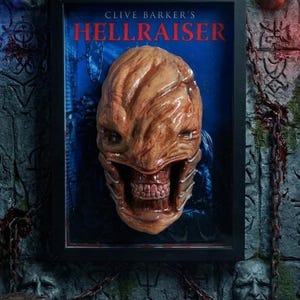 May include: A framed display featuring a sculpted, flesh-toned head with a wide, toothy mouth and textured skin. The background is deep blue with the text CLIVE BARKER'S HELLRAISER in red. The frame is black and rectangular, set against a stone wall.
