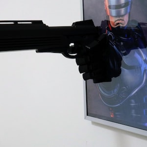 Pu&ograve; includere: Una pistola di scena nera si estende da un'immagine incorniciata di un personaggio di fantascienza. La pistola &egrave; tenuta da una mano guantata di nero. L'immagine &egrave; in una cornice argentata e montata su un muro bianco.