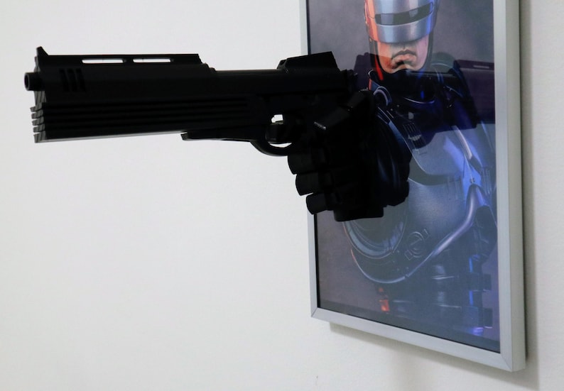 Pu&ograve; includere: Una pistola di scena nera si estende da un'immagine incorniciata di un personaggio di fantascienza. La pistola &egrave; tenuta da una mano guantata di nero. L'immagine &egrave; in una cornice argentata e montata su un muro bianco.