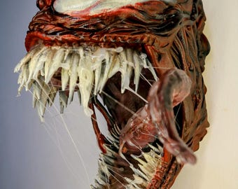 Può includere: Scultura tridimensionale dettagliata di una testa mostruosa, prevalentemente rossa e marrone con accenti bianchi. La bocca aperta rivela denti affilati biancastri e una lunga lingua rossa. La scultura ha un aspetto strutturato e organico.