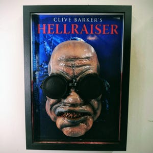 May include: Framed artwork depicting a disturbing, pale face with large, round black goggles and a grimacing mouth. The background shows the title "Clive Barker's Hellraiser" in red letters against a blue backdrop. The artwork is in a black frame.